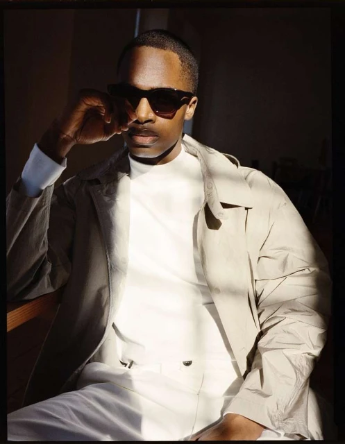 Portrait of Black man in sunglasses and trench coat in subdued lighting. He is seated, wearing a white turtleneck and pants.