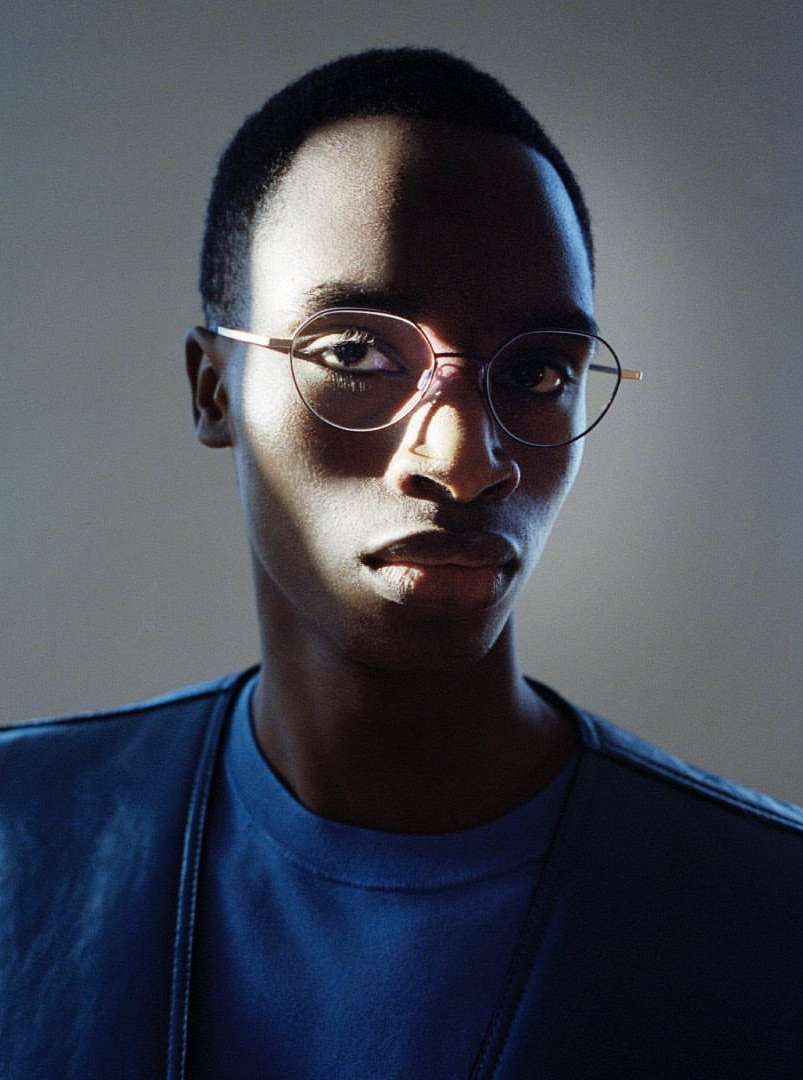 Headshot of adult Black male wearing glasses and a blue shirt. The image is cropped tightly and lit with bright light and dark shadow.