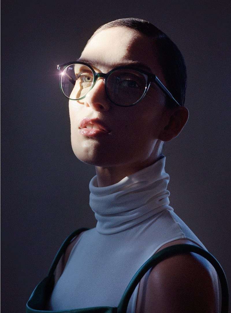 Adult female wearing glasses and a white turtleneck with green dress straps. Light shines across the subject's face, illuminating the cheekbone.
