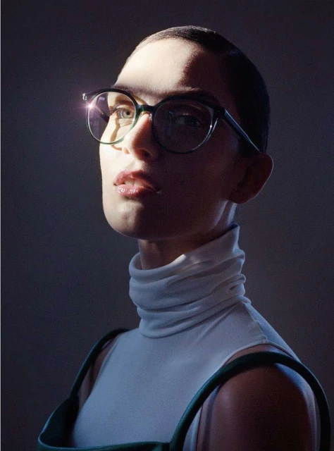 Adult female wearing glasses and a white turtleneck with green dress straps. Light shines across the subject's face, illuminating the cheekbone.