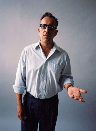 Man wearing sunglasses and light blue button-down shirt gesturing against grey seamless backdrop
