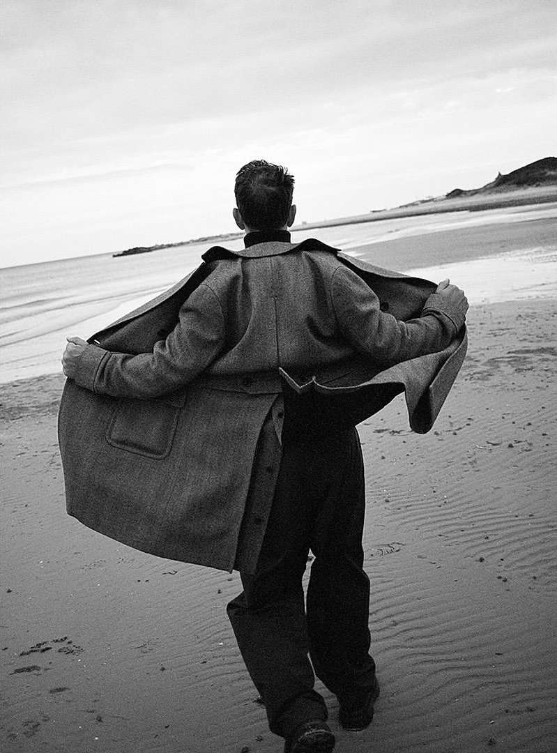 Man wearing a long wool coat walking along a sandy beach with the coat billowing open in the wind