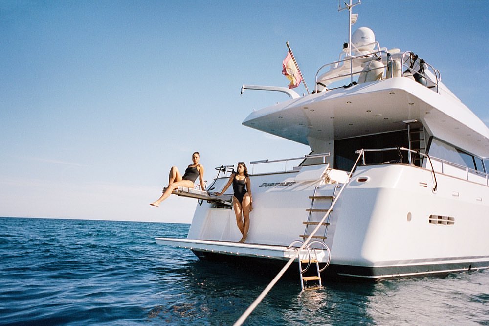 Two women in swimsuits pose on the stern of a large white yacht at sea under a clear blue sky, capturing a luxury lifestyle scene.