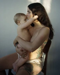 Mother kissing her toddler child while seated on a wooden chair against a plain wall in a portrait by Petra Kleis.