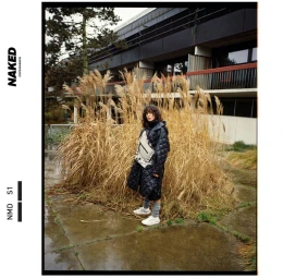 Model wearing a black quilted coat and white sneakers standing outdoors near tall dry grass in front of a modern building.