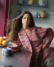 Woman in patterned red kimono leaning on a dark counter holding a blue mug, with bowls of citrus fruit and shelves of pottery behind.