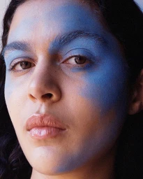 Close-up portrait of a woman with bold blue paint makeup across her forehead, brow, and cheek, photographed by Petra Kleis.