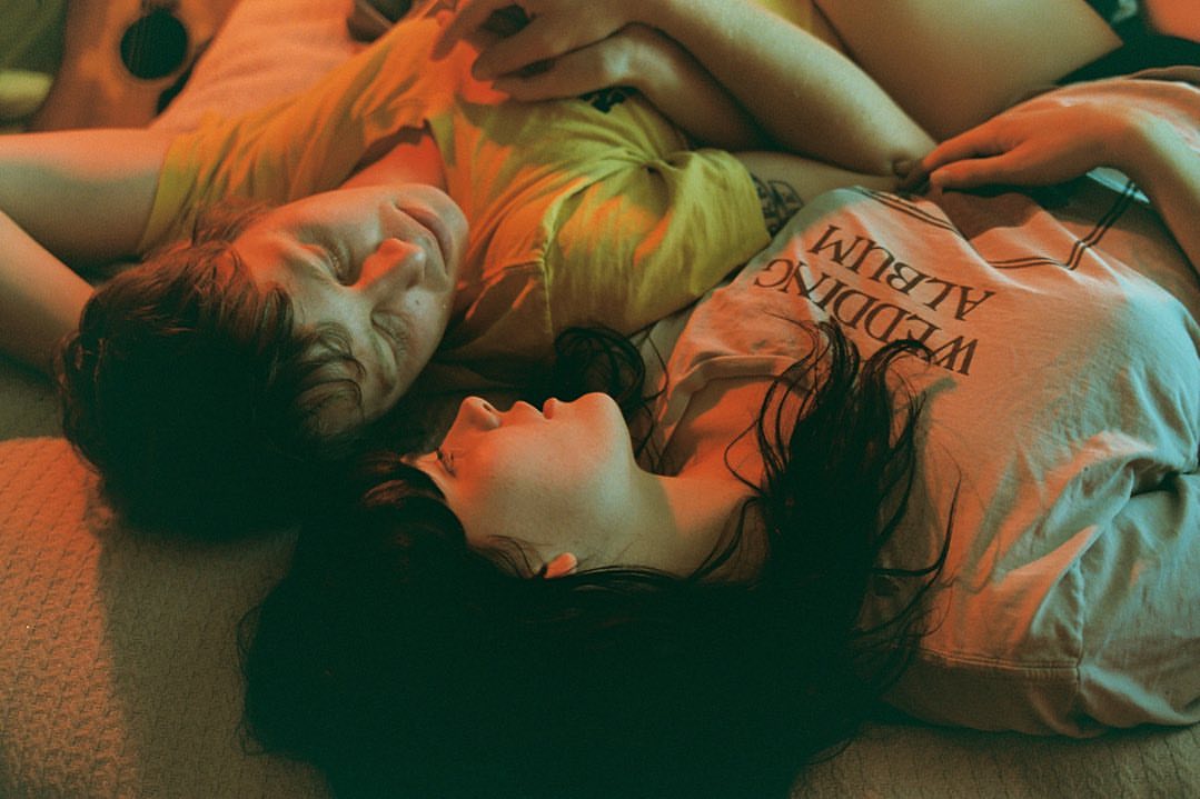 Two young adults in casual apparel rest together on a bed indoors, soft, warm lighting, and a close-up shot capturing a moment of relaxation.