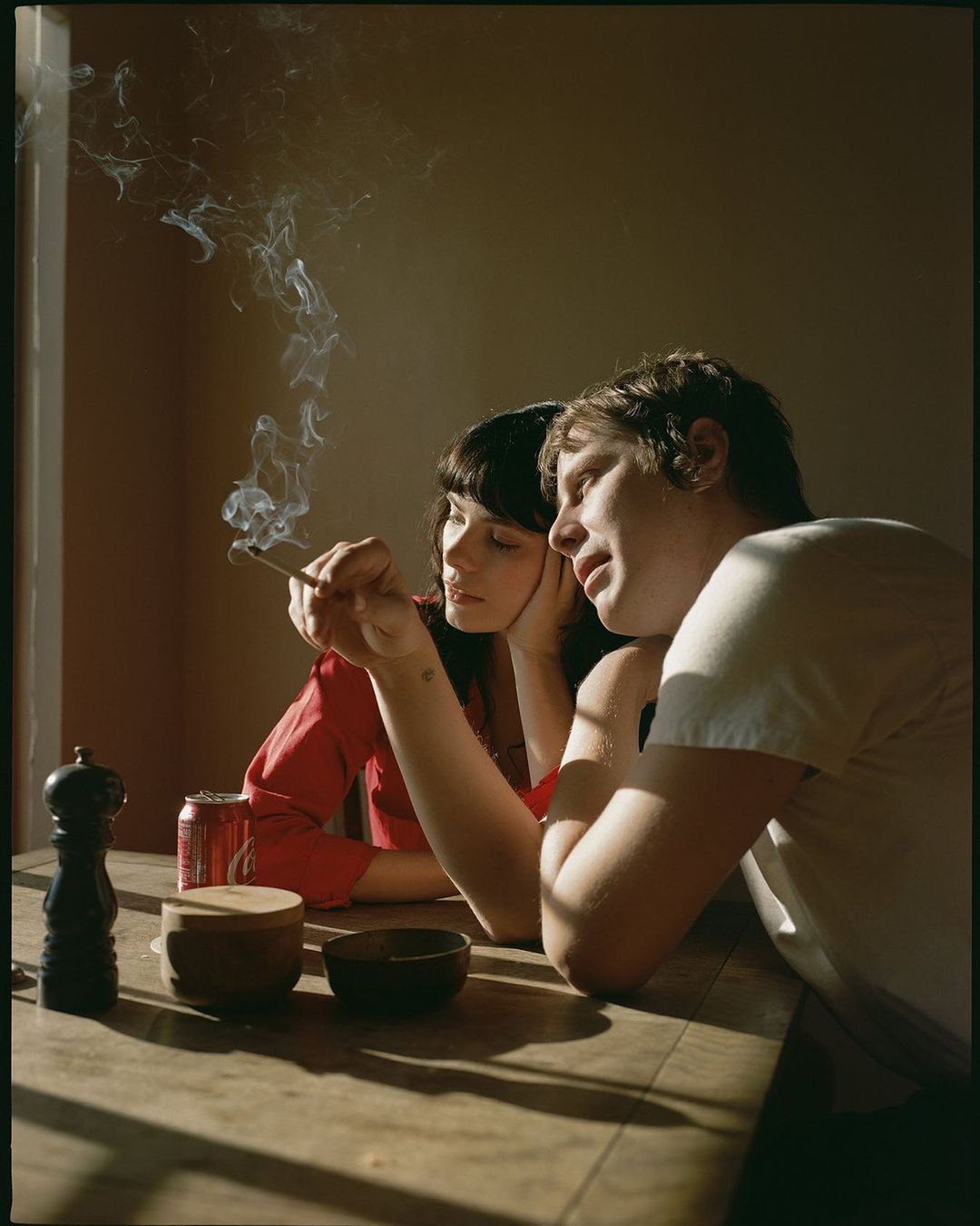 Indoor portrait of a couple seated at a wooden table, one smoking, illuminated by window light, accompanied by wooden bowls and a beverage can.