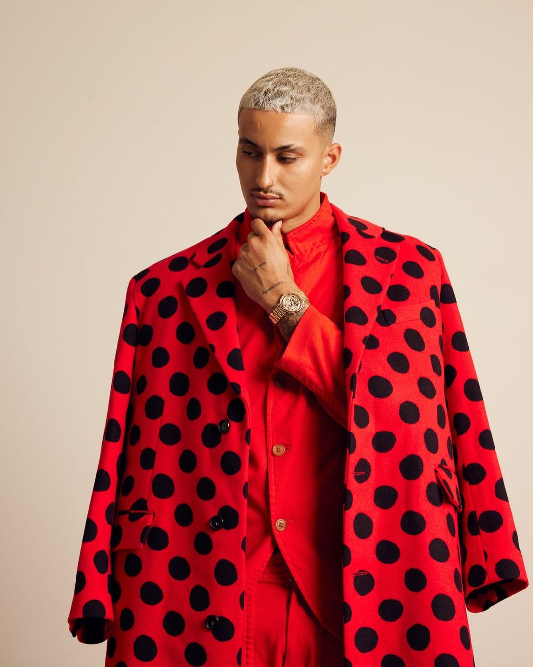 Stylish man poses in a red suit jacket with black polka dots, paired with a matching red shirt, in a neutral studio setting.