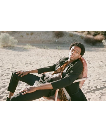 Black man seated in a weathered chair on a sandy beach wearing a black blazer and patterned scarf