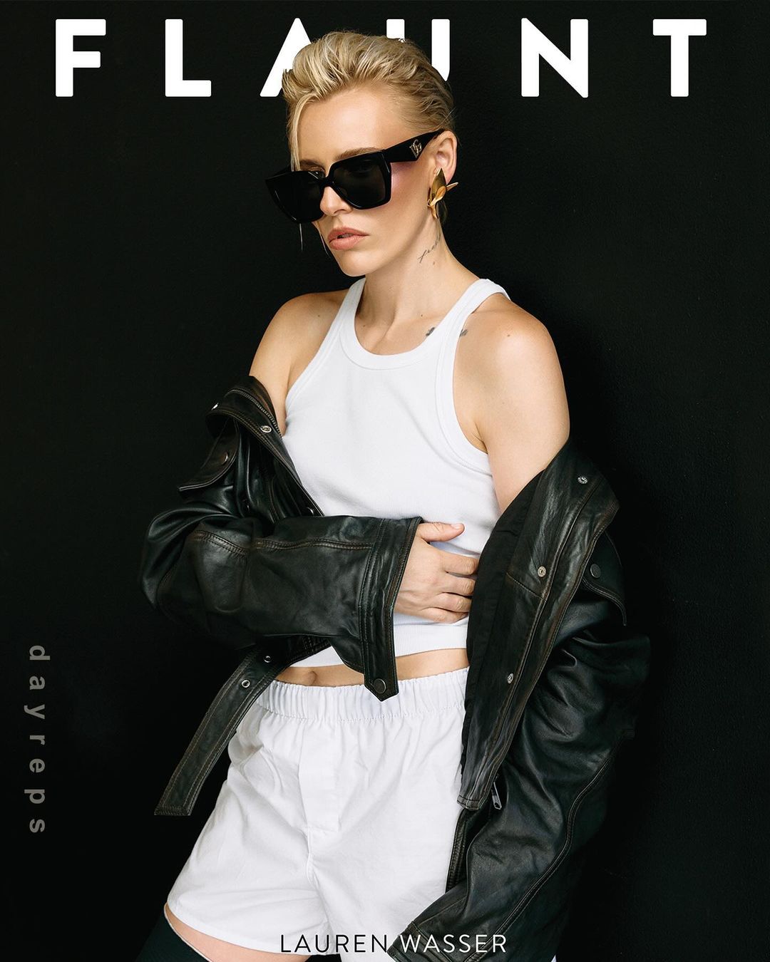 Portrait of a stylish woman with short blonde hair wearing sunglasses, a white tank top, and a black jacket against a black background.