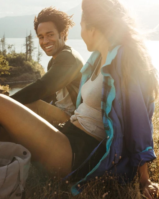 Smiling man and woman relaxing outdoors on grassy hillside with lake and mountains in background on sunny day in daylight.