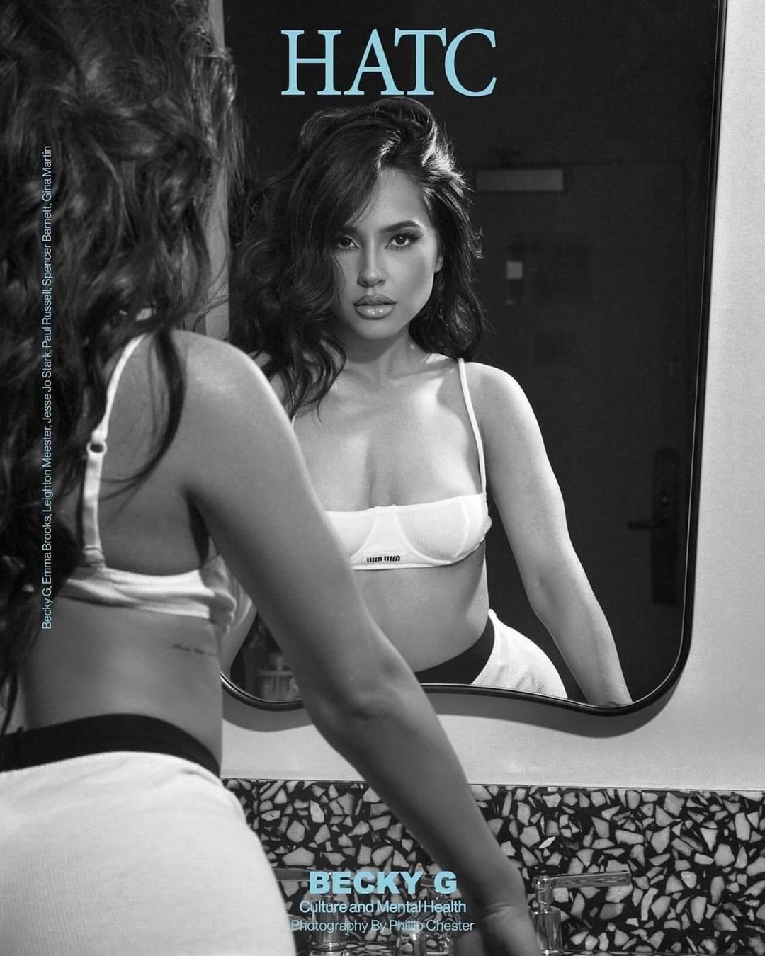 Brunette woman in a white bra and black and white shorts looks in a mirror in an indoor setting. The image is black and white.