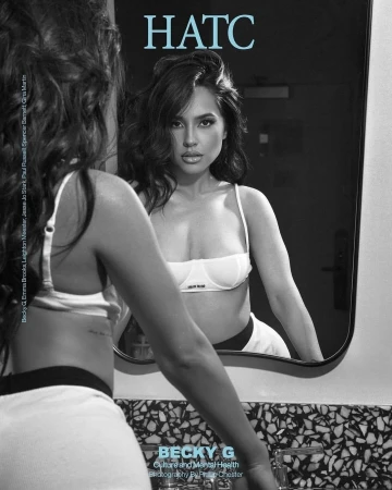 Brunette woman in a white bra and black and white shorts looks in a mirror in an indoor setting. The image is black and white.