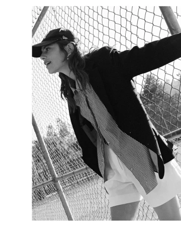 Black and white portrait of a woman wearing a baseball cap, dark blazer, striped shirt, and light shorts against a chain-link fence.