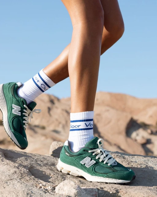 Close-up of New Balance green running shoes with white socks on a light brown textured surface against a blue sky backdrop.
