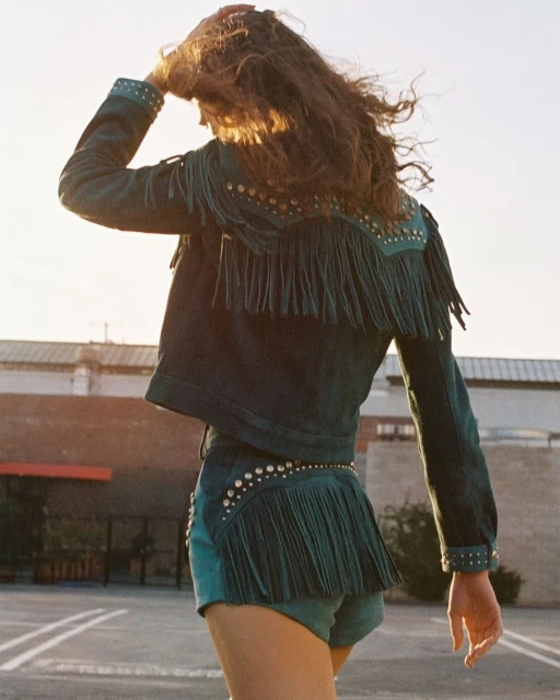 A person wearing a blue fringe jacket and shorts stands outdoors on a sunny day with city buildings in the background in a fashion shot.