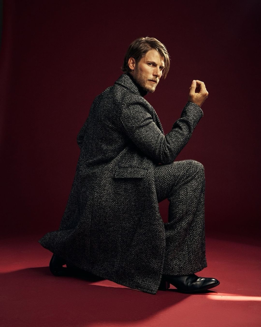 A man with blond hair poses in a grey herringbone coat and matching suit pants while kneeling on a red seamless backdrop looking back.