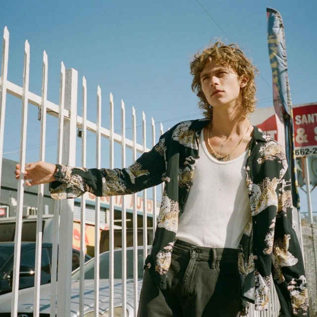 A young adult male stands near a white fence in an urban setting with a car repair shop sign visible in the background on a sunny day.