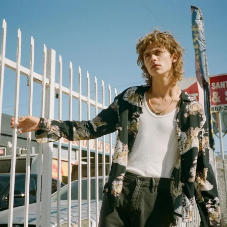 A young adult male stands near a white fence in an urban setting with a car repair shop sign visible in the background on a sunny day.