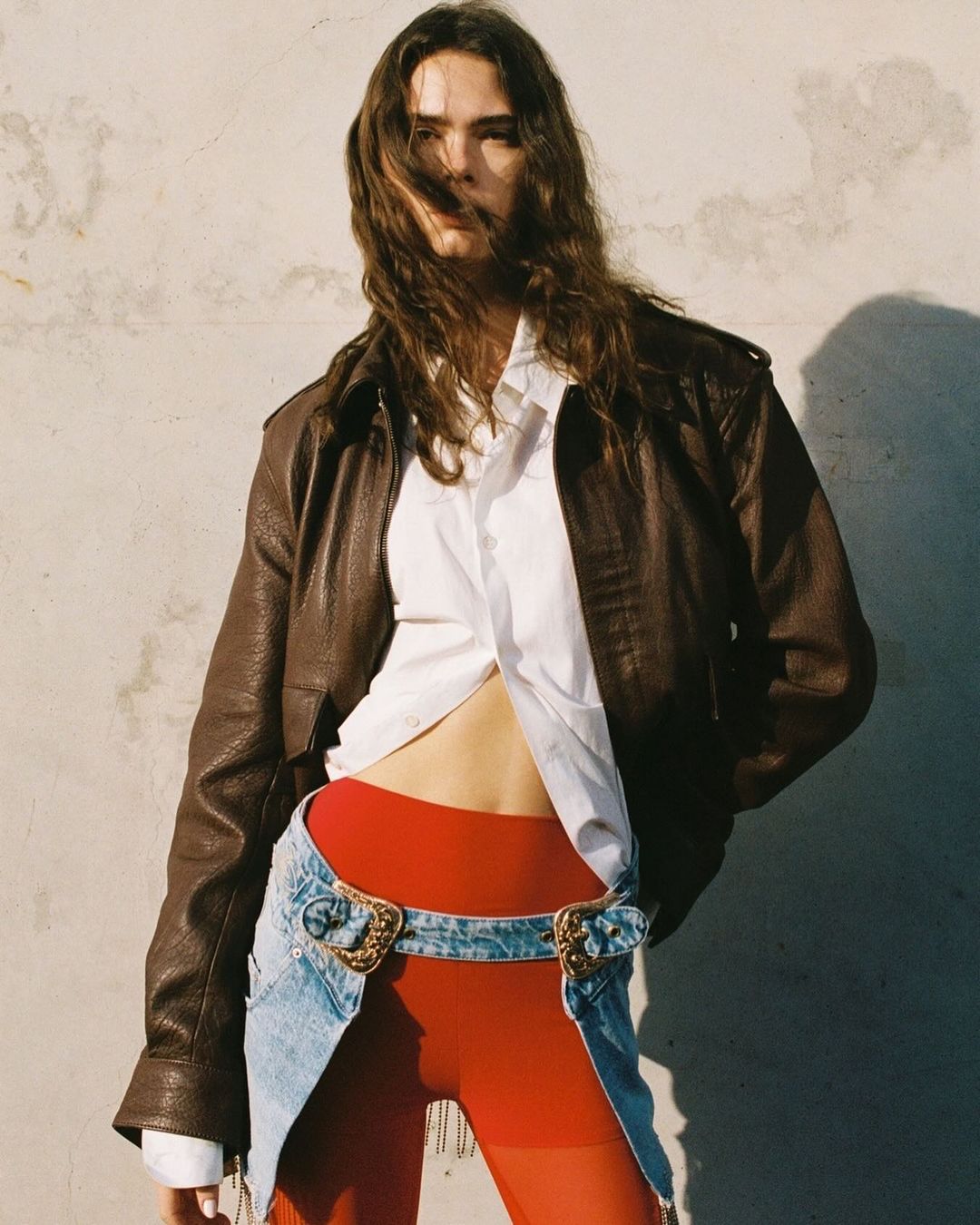 A female fashion model poses outdoors wearing a brown leather jacket, white top, denim belt-skirt, and red leggings against a concrete wall.