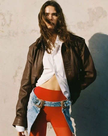 A female fashion model poses outdoors wearing a brown leather jacket, white top, denim belt-skirt, and red leggings against a concrete wall.