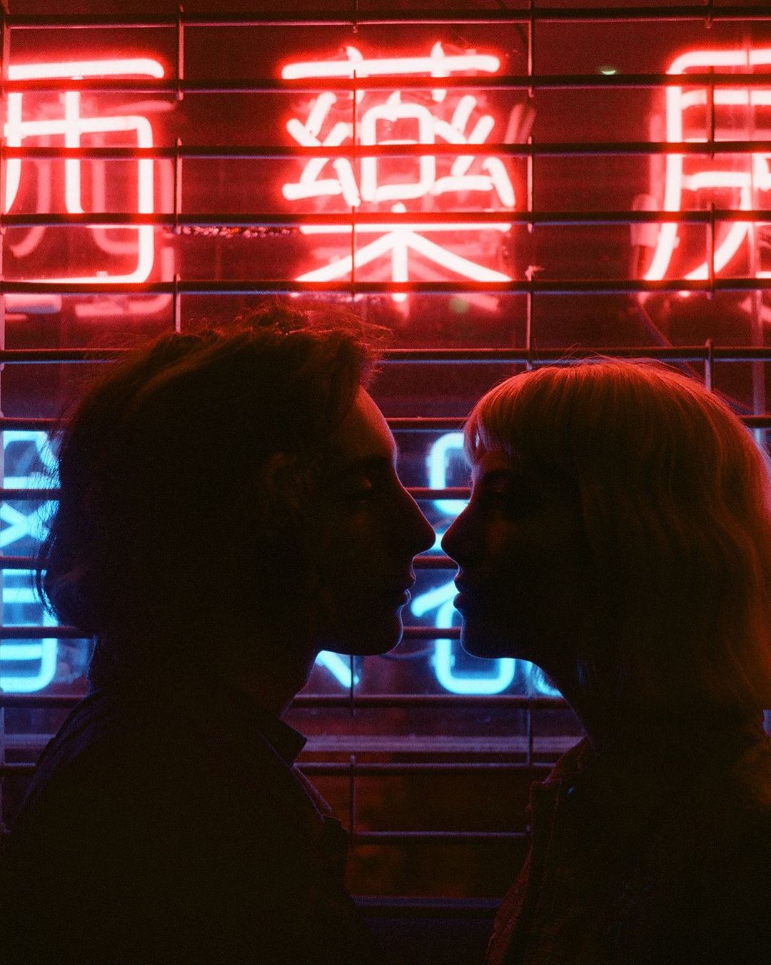 Silhouetted couple facing each other in front of neon sign with Asian characters. Dark mood lighting and close framing.
