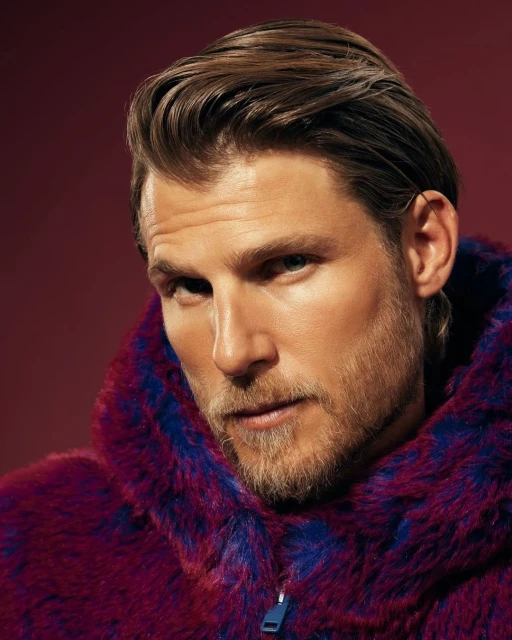 Close-up portrait of a handsome, blond, bearded male model with blue eyes wearing a purple and blue faux fur coat against a red backdrop.