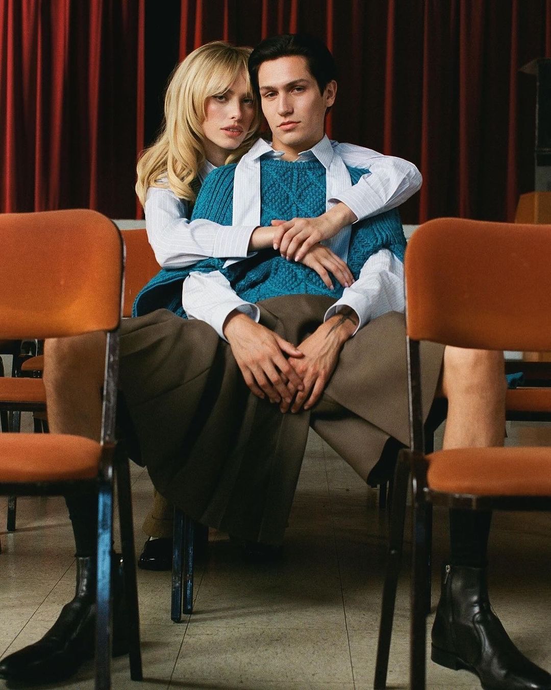 Editorial fashion shot of a couple in an auditorium. The woman has her arms around the man as they sit in chairs in front of red curtains.