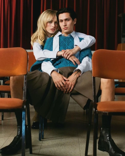 Editorial fashion shot of a couple in an auditorium. The woman has her arms around the man as they sit in chairs in front of red curtains.