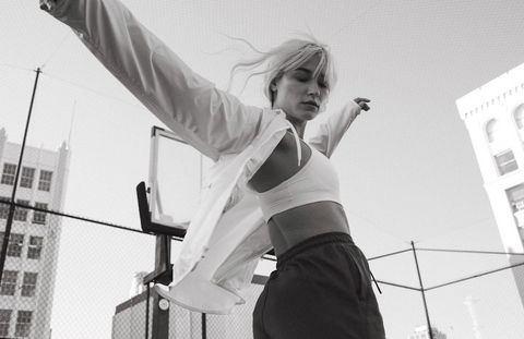 Athletic woman in sports bra and track pants poses with arms outstretched against a city building backdrop in monochrome.
