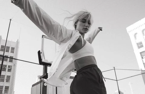 Athletic woman in sports bra and track pants poses with arms outstretched against a city building backdrop in monochrome. 