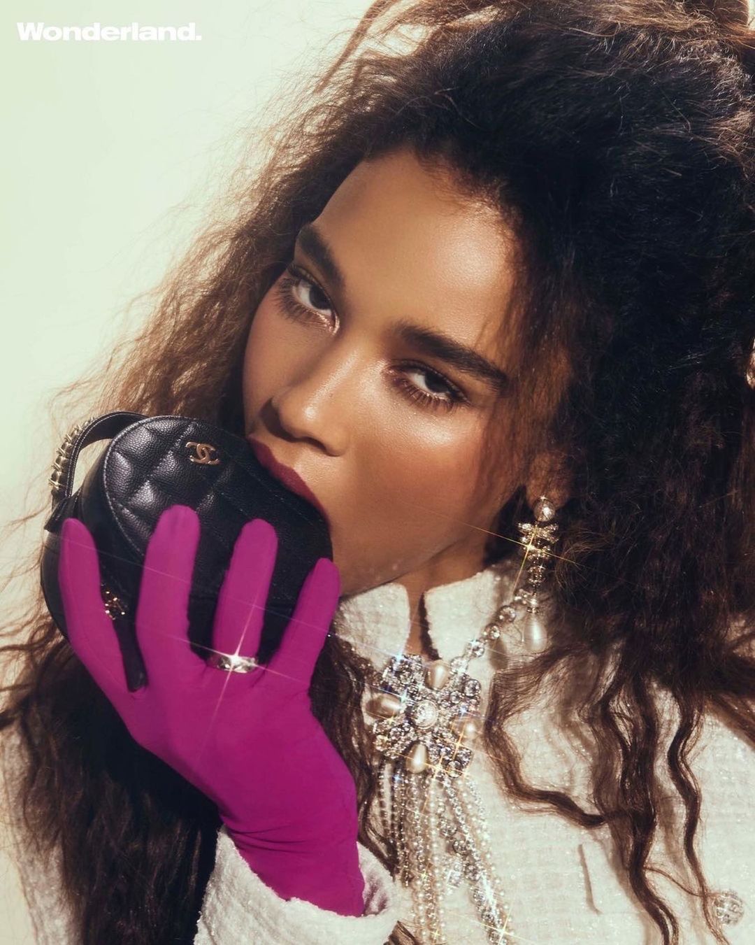 Close-up of a female fashion model with curly brown hair wearing pearls, jewelry, and a jacket with a pink glove holding a handbag.