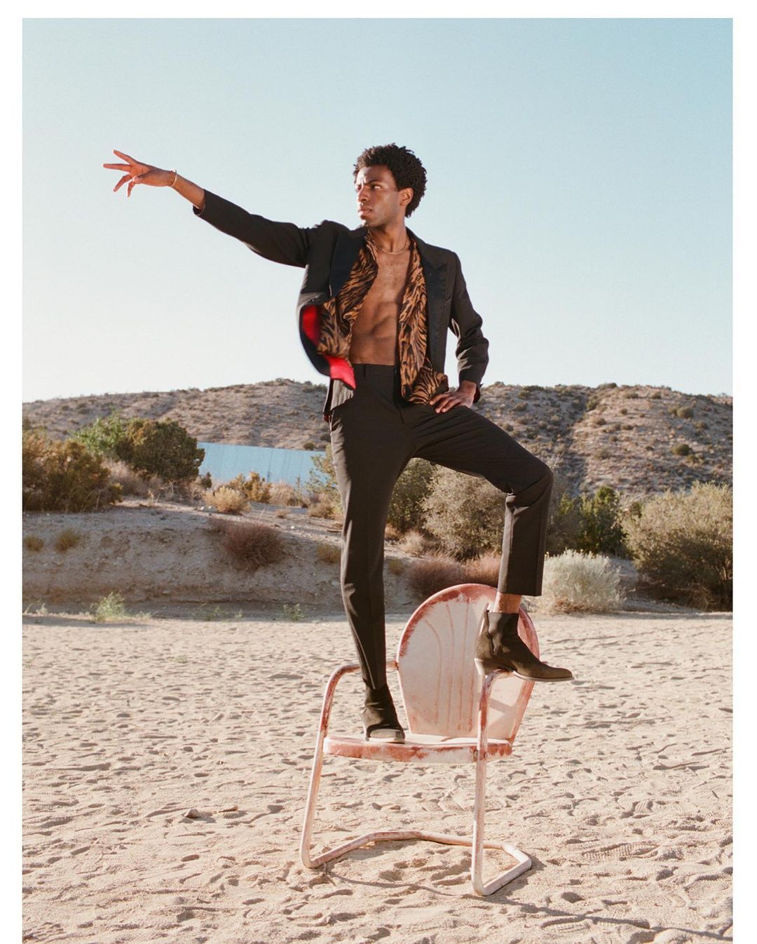 Black man in dark suit and patterned open shirt standing with one foot on a metal chair in a desert landscape