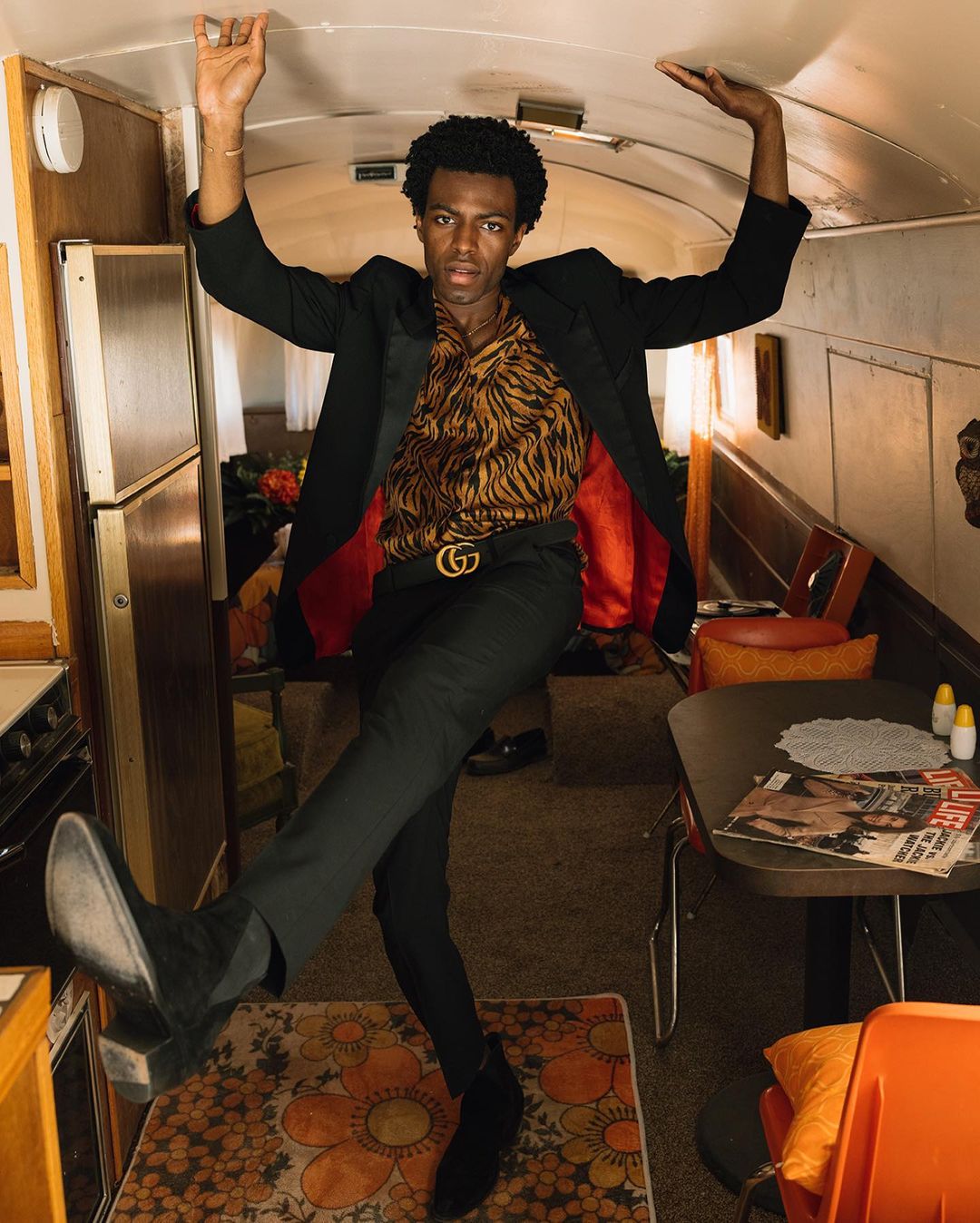 Stylish African-American man in a blazer, patterned shirt, and black pants poses inside of a vintage recreational vehicle with a floral rug.