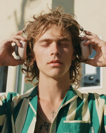 Young man with curly blonde hair and closed eyes wearing headphones and a striped shirt by Phil And Sara