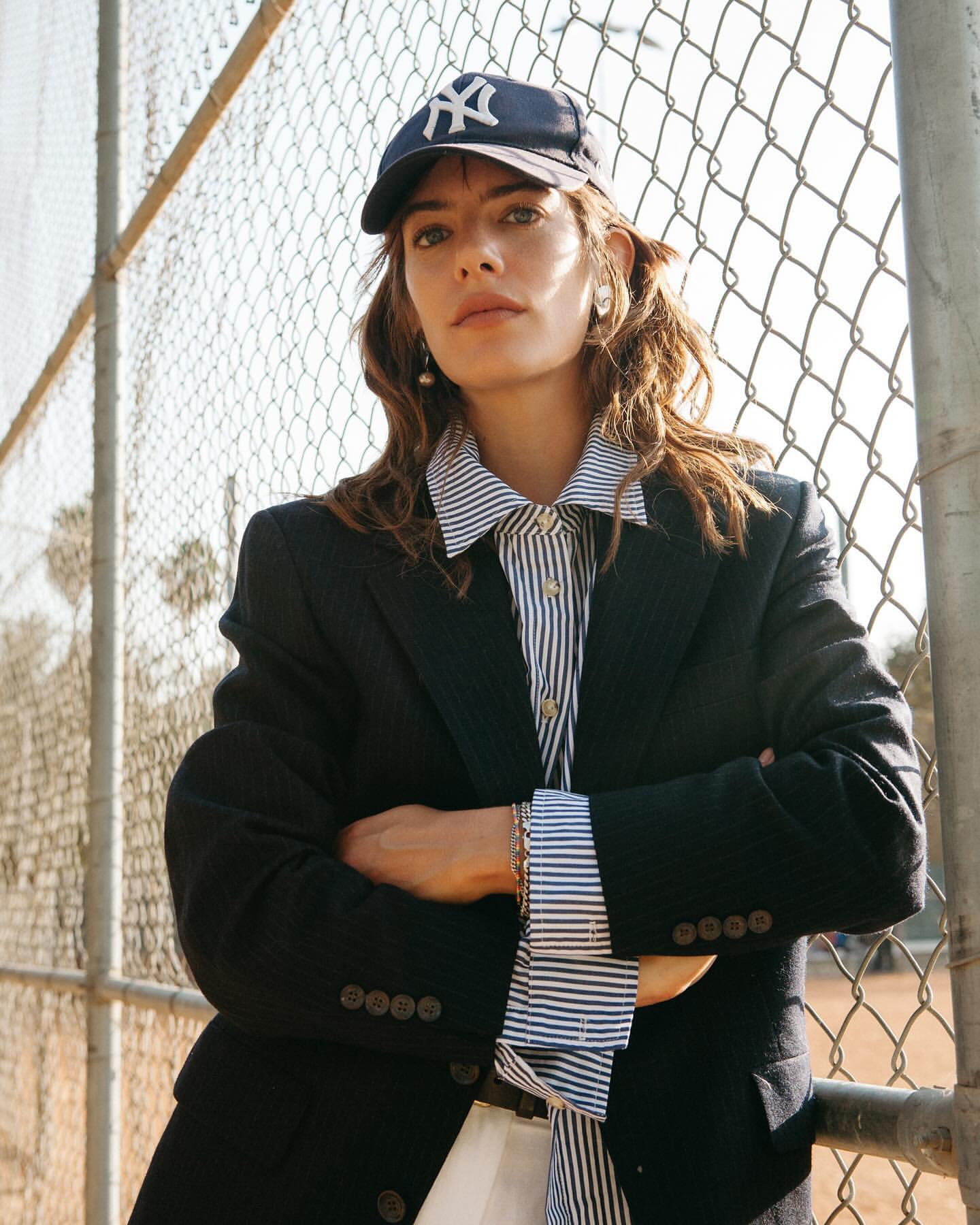 Woman wearing a dark baseball cap and blazer with arms crossed against a chain link fence outdoors in Los Angeles.