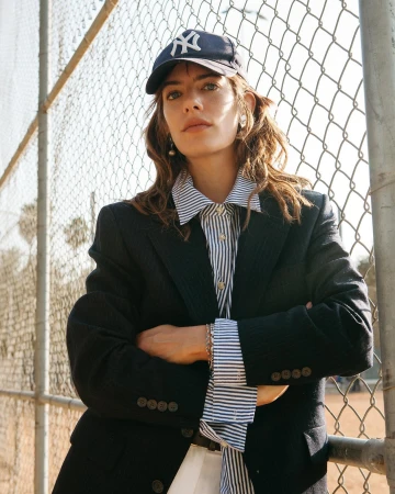 Woman wearing a dark baseball cap and blazer with arms crossed against a chain link fence outdoors in Los Angeles.