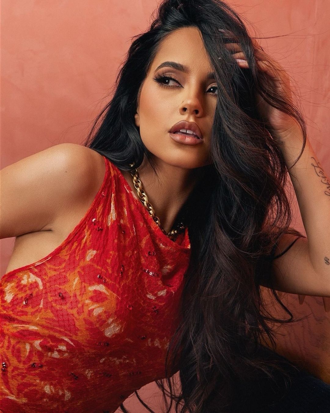 Close-up portrait of a woman with long dark hair, wearing a red patterned top and gold chain necklace, posing against a peach background.