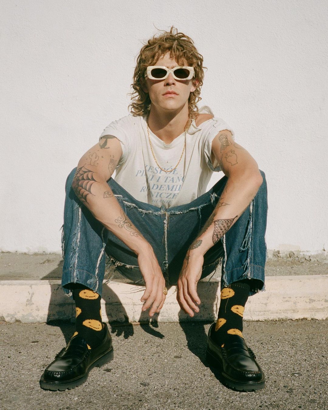 Young adult with curly hair, sunglasses, and tattoos posing on a concrete step in jeans, t-shirt, and loafers with smiley-face socks.