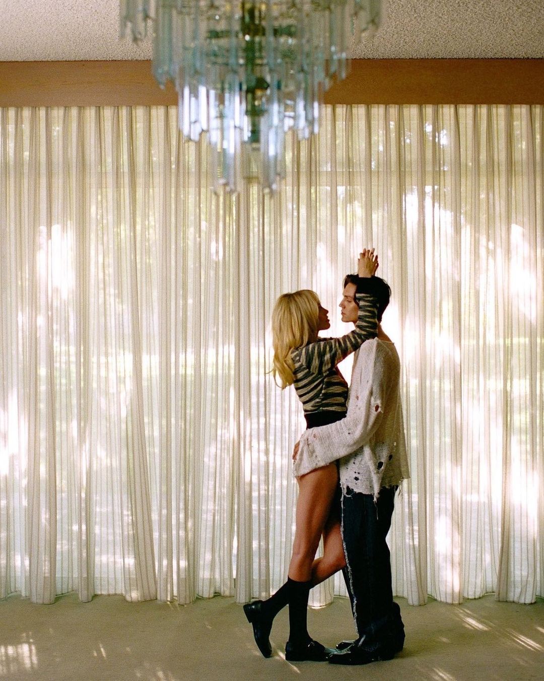 Two people embracing closely in a residential room with sheer white curtains and a hanging crystal chandelier.