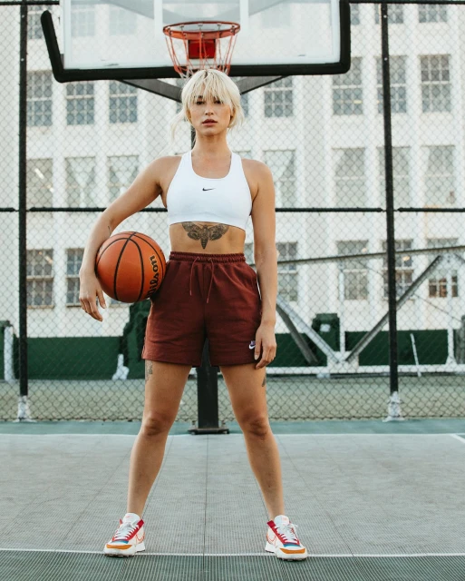 Young woman with short blonde hair in sportswear poses with a basketball on an outdoor court in front of a tall building.