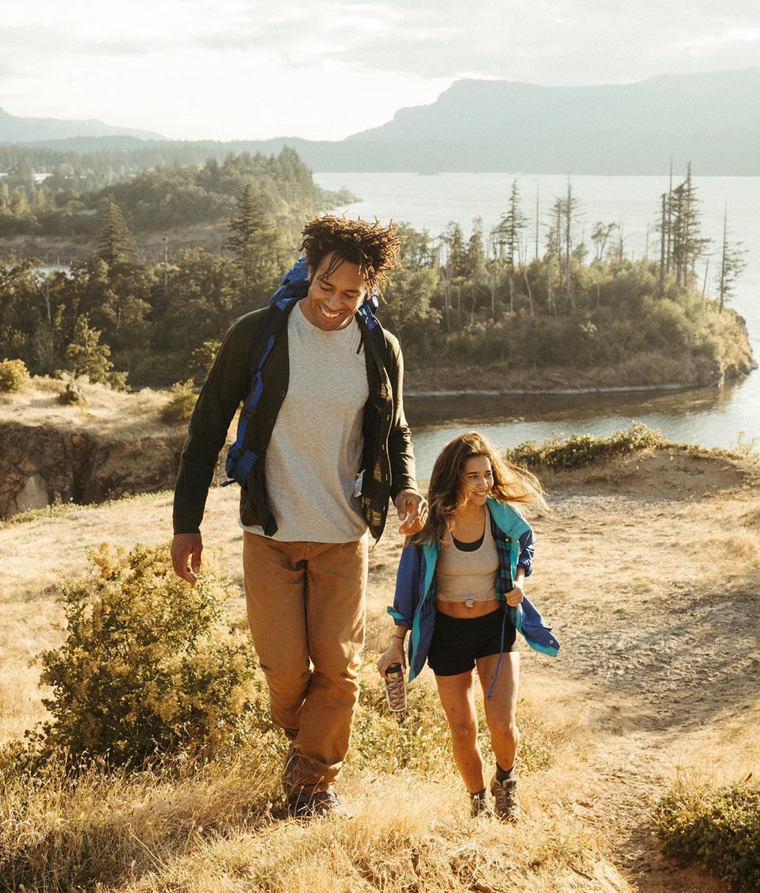 Couple hikes a grassy hill overlooking a lake on a sunny day. The man has a backpack, and they are holding hands on the trail.