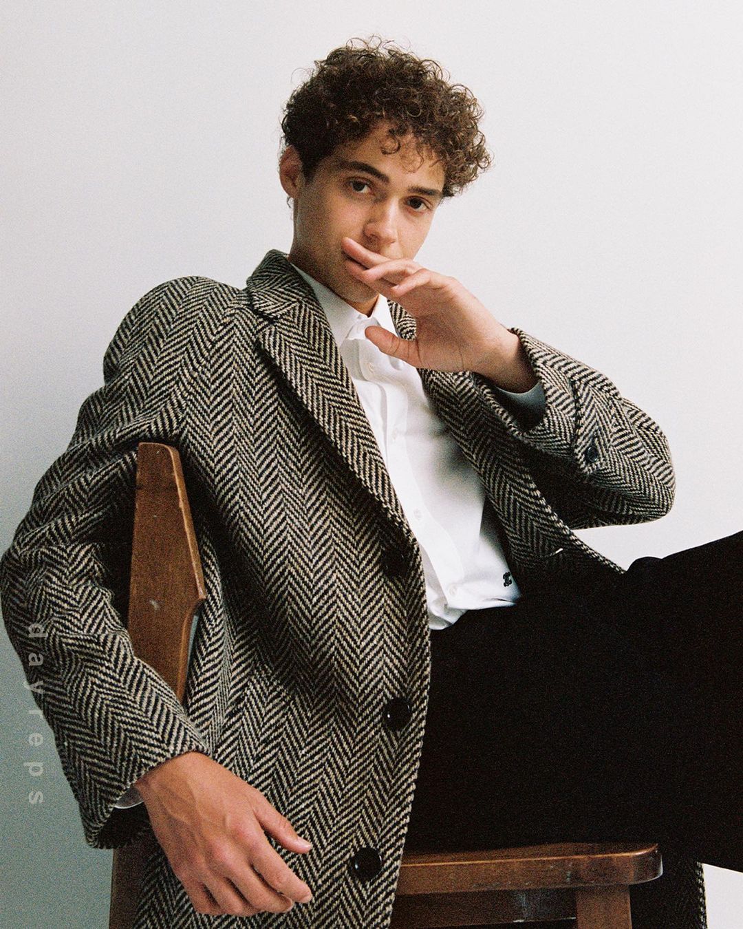 Portrait of a curly-haired man in a patterned coat sitting on a chair in front of a neutral background, stylish and confident.