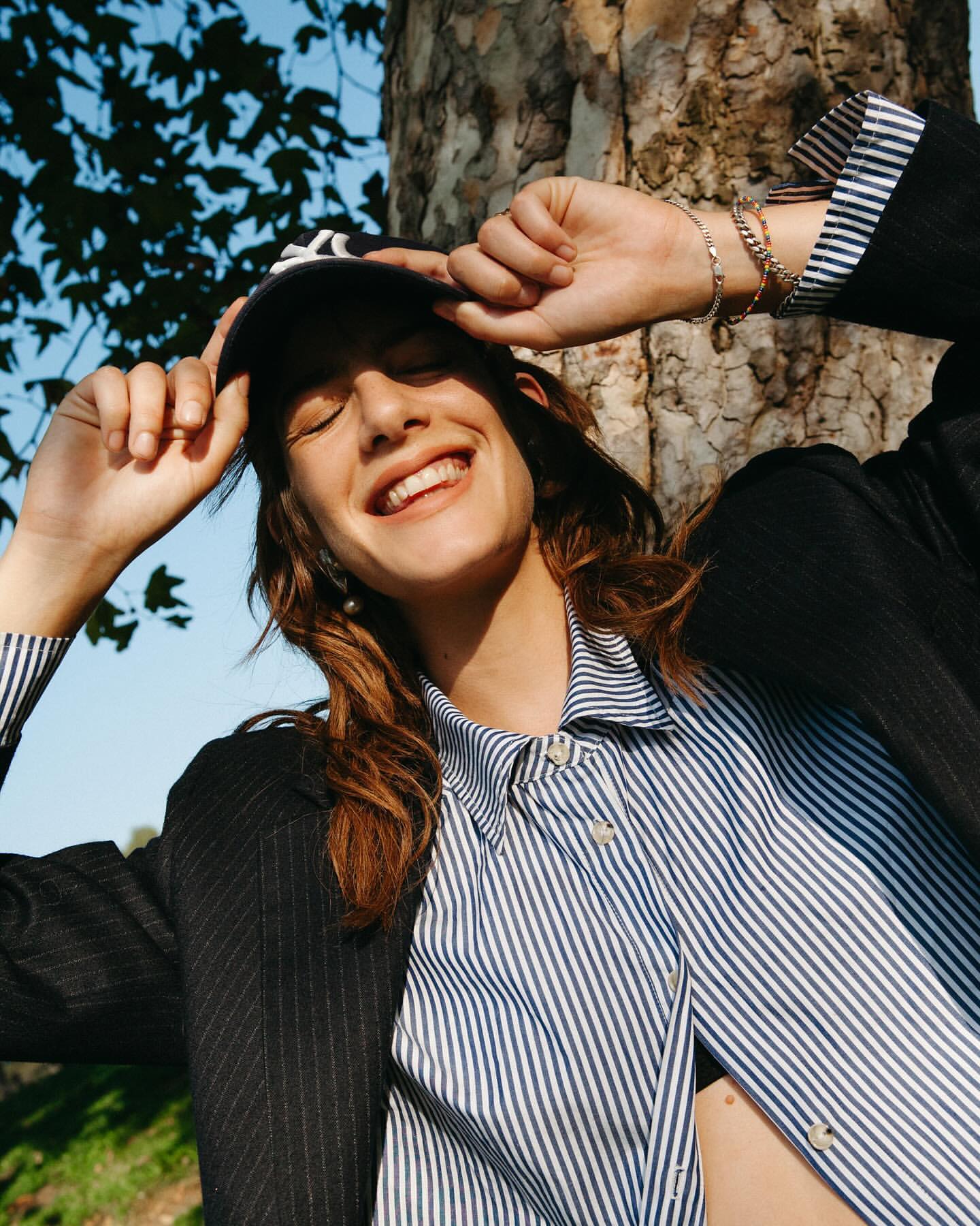 Young woman smiles outdoors wearing a baseball cap, striped shirt, and blazer, enjoying natural light and shadows under tree branches.