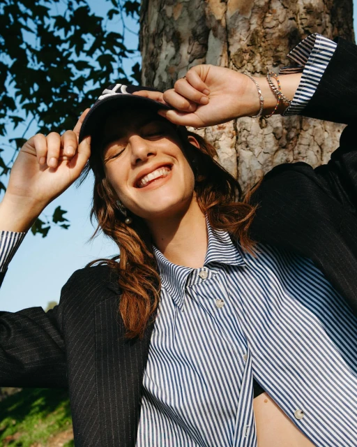 Young woman smiles outdoors wearing a baseball cap, striped shirt, and blazer, enjoying natural light and shadows under tree branches.