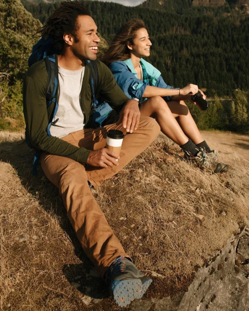 Two hikers, an adult Black male holding a cup and an adult white female holding binoculars, sit on a hillside overlooking a mountain forest.