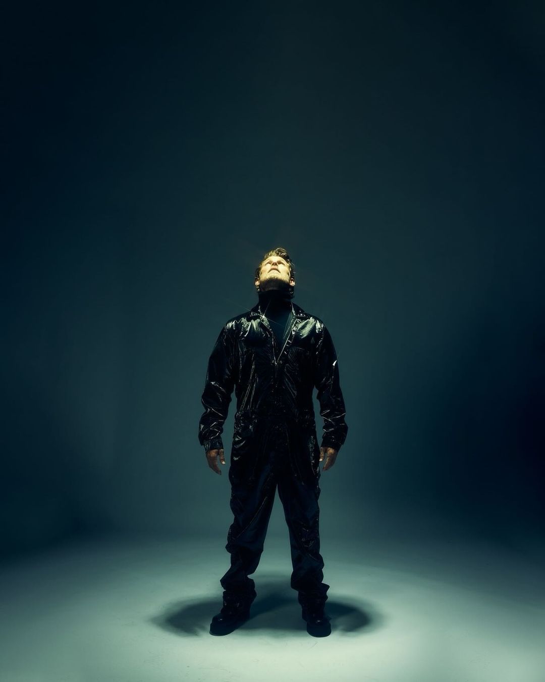 Man in a black jumpsuit gazes upwards in a dramatic pose against a dark background, highlighted by strong directional lighting from above.