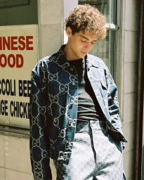 A young man with curly hair wearing a blue monogram denim jacket standing outside a building in Los Angeles.